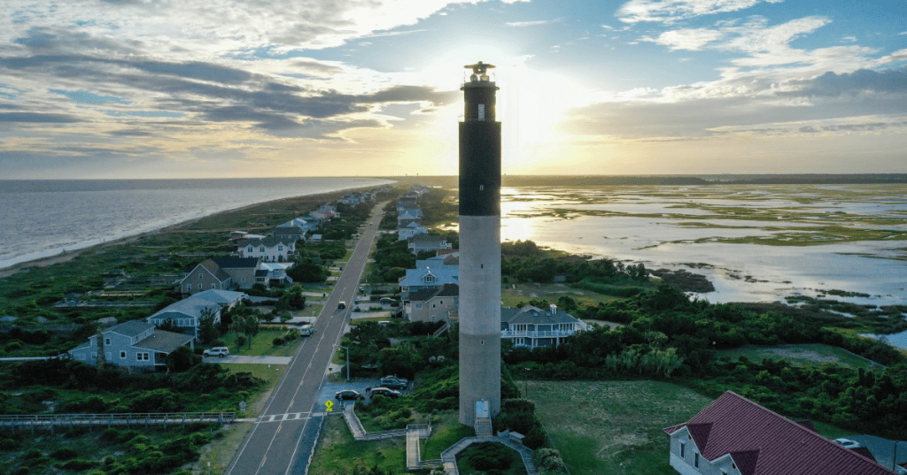 The sun setting behind the striped Oak Island Lighthouse on a cloudy day with houses and marshland below it.