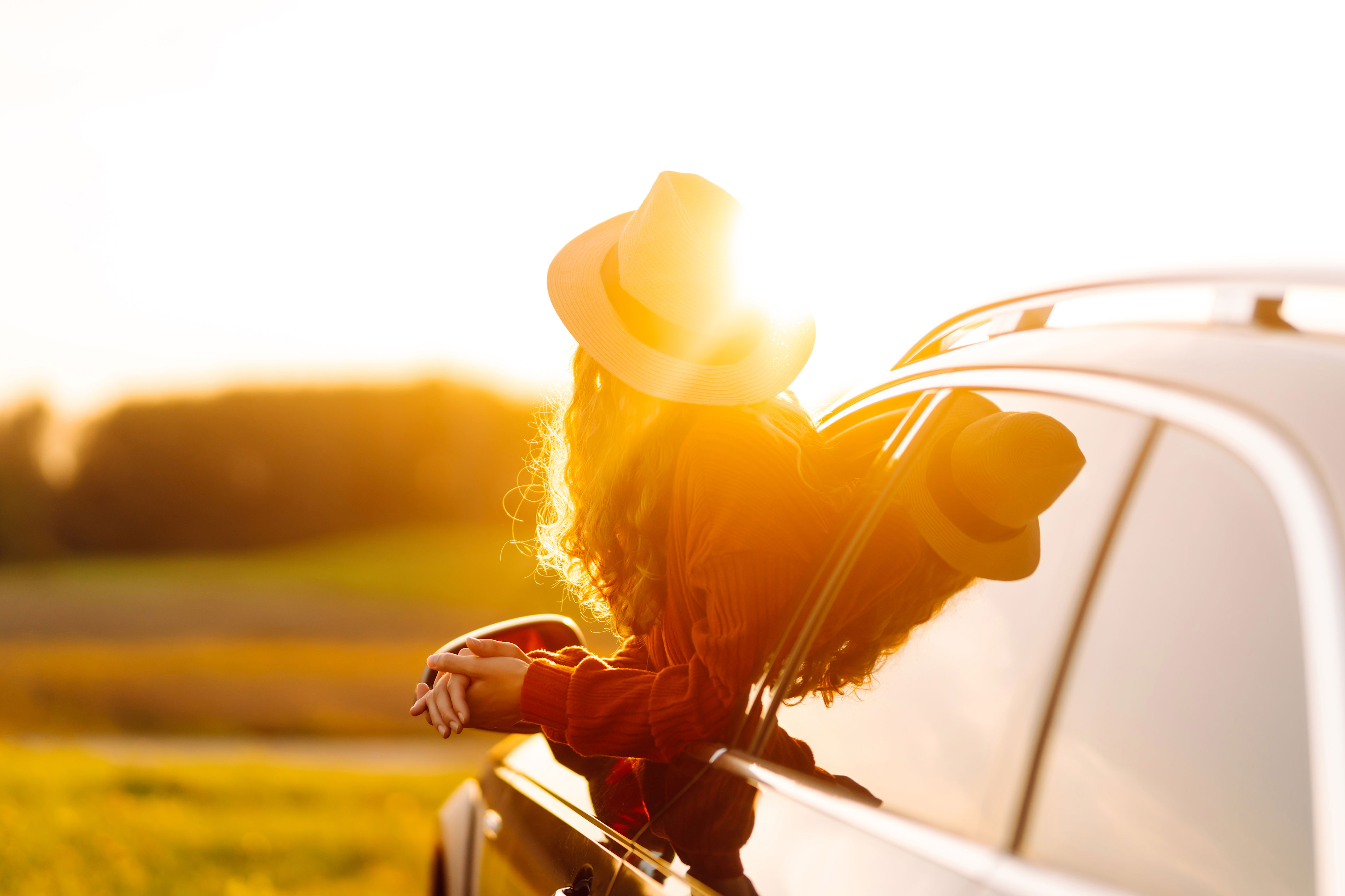 Person in a hat leans out of a car window, enjoying a sunset view in a rural setting. Their face is not visible