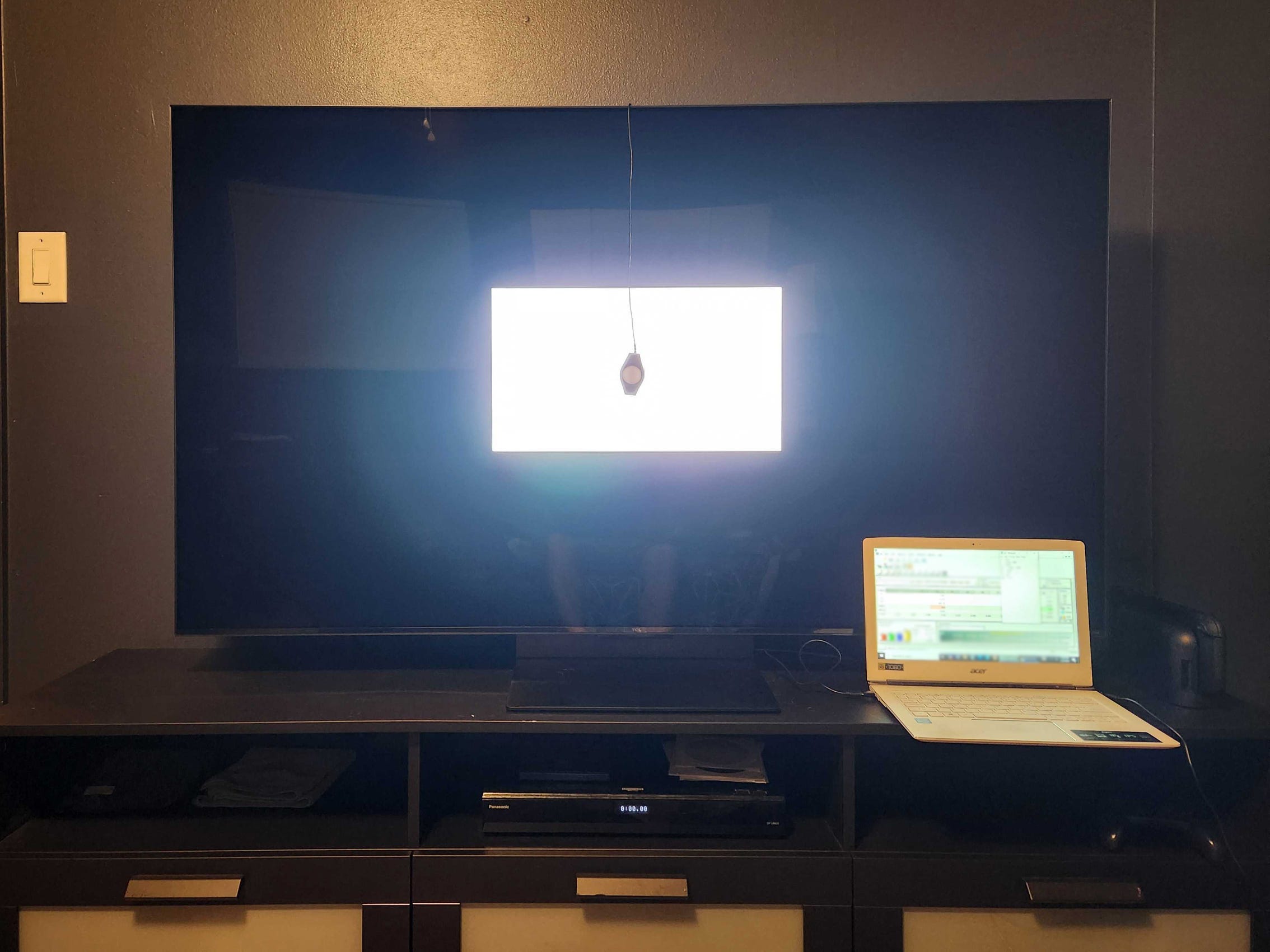 A TCL QM7K TV on a media console with a colorimeter placed on a white box displayed on the screen.