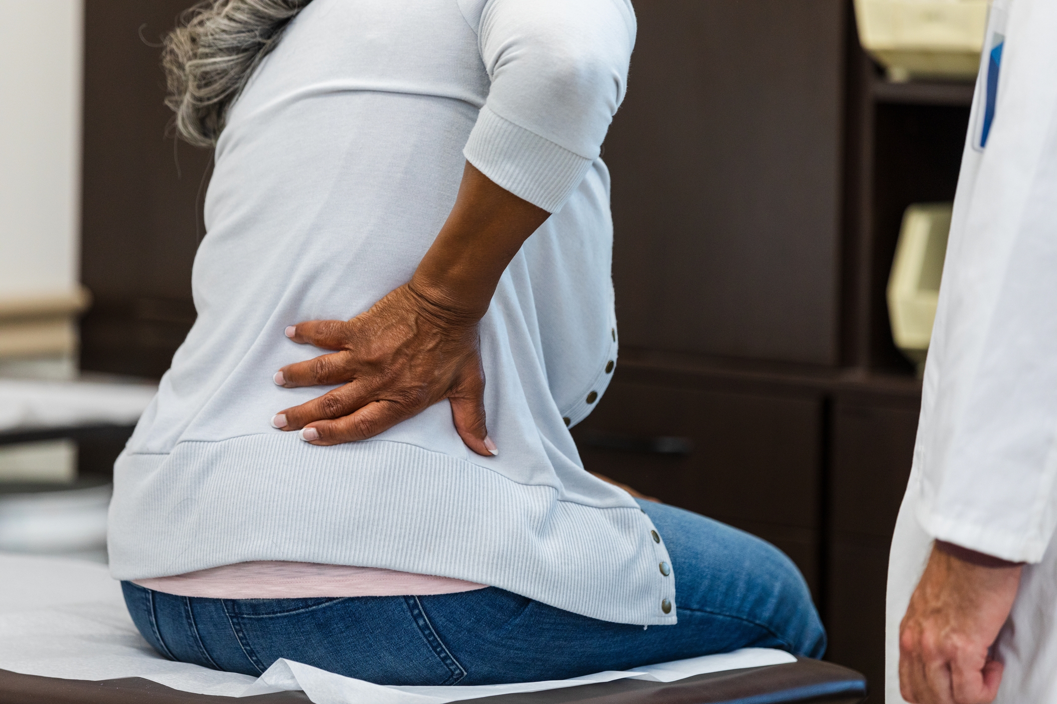 Person sitting on an exam table holding their lower back, expressing discomfort, with a healthcare professional nearby