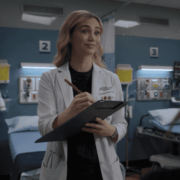 Medical professional smiling while holding a clipboard in a hospital room with patient beds in the background