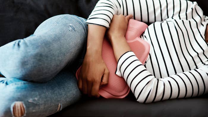 Person in striped shirt and jeans lies on a couch holding a hot water bottle to their abdomen, suggesting comfort or relief