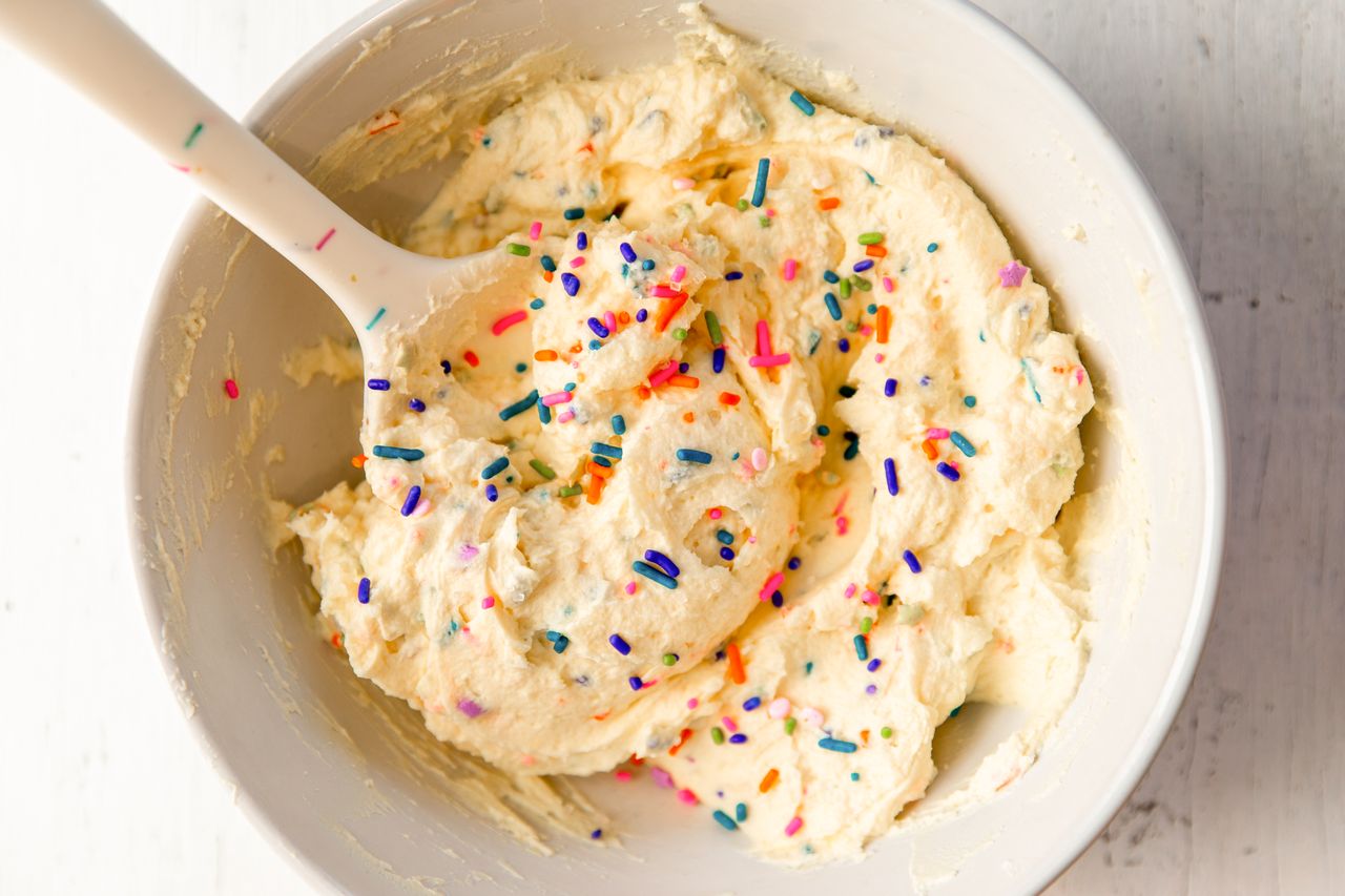 A bowl of yogurt frosting topped with sprinkles, stirred with a spoon