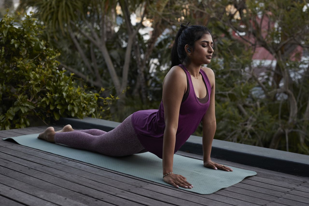 Full length of young woman wearing in-ear headphones practicing cobra pose yoga with eyes closed on exercise mat at patio in back yard during sunset