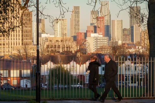 Commuters pass a London skyline of high-rise towers that are seen through bare branches of ash trees bordering Ruskin Park on a cold winter's morning in South London