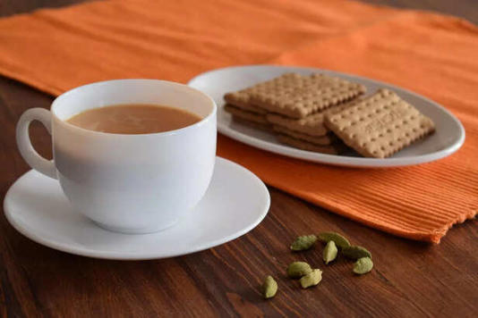 a image showing a cup of tea , buiscuit and some cardamom pods