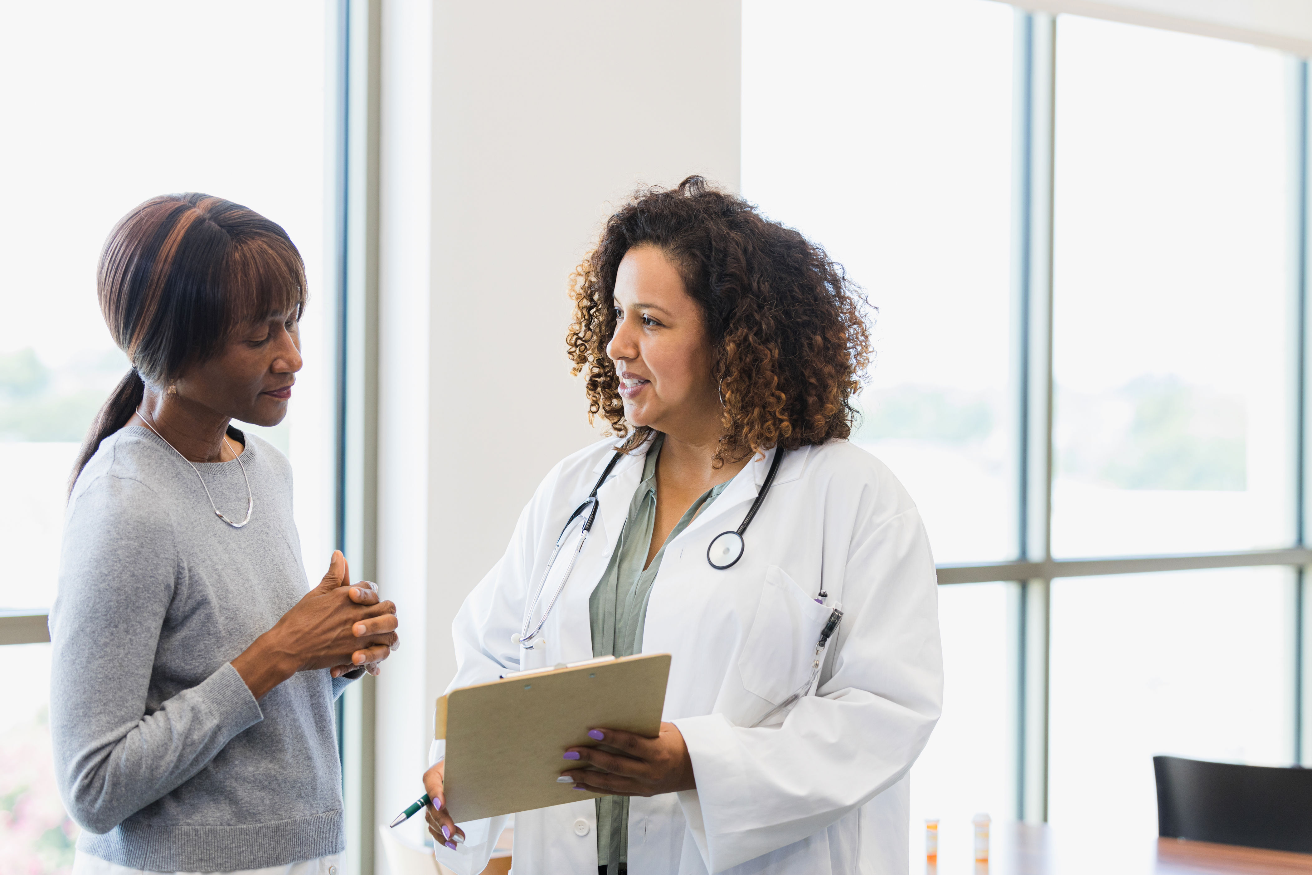 Doctor and patient discussing health in a bright office. Doctor holds a clipboard and wears a stethoscope. Both appear engaged in conversation