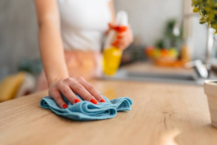 Person cleaning a wooden surface with a cloth, holding a spray bottle. Focus on hand and surface, emphasizing household chores related to work and money