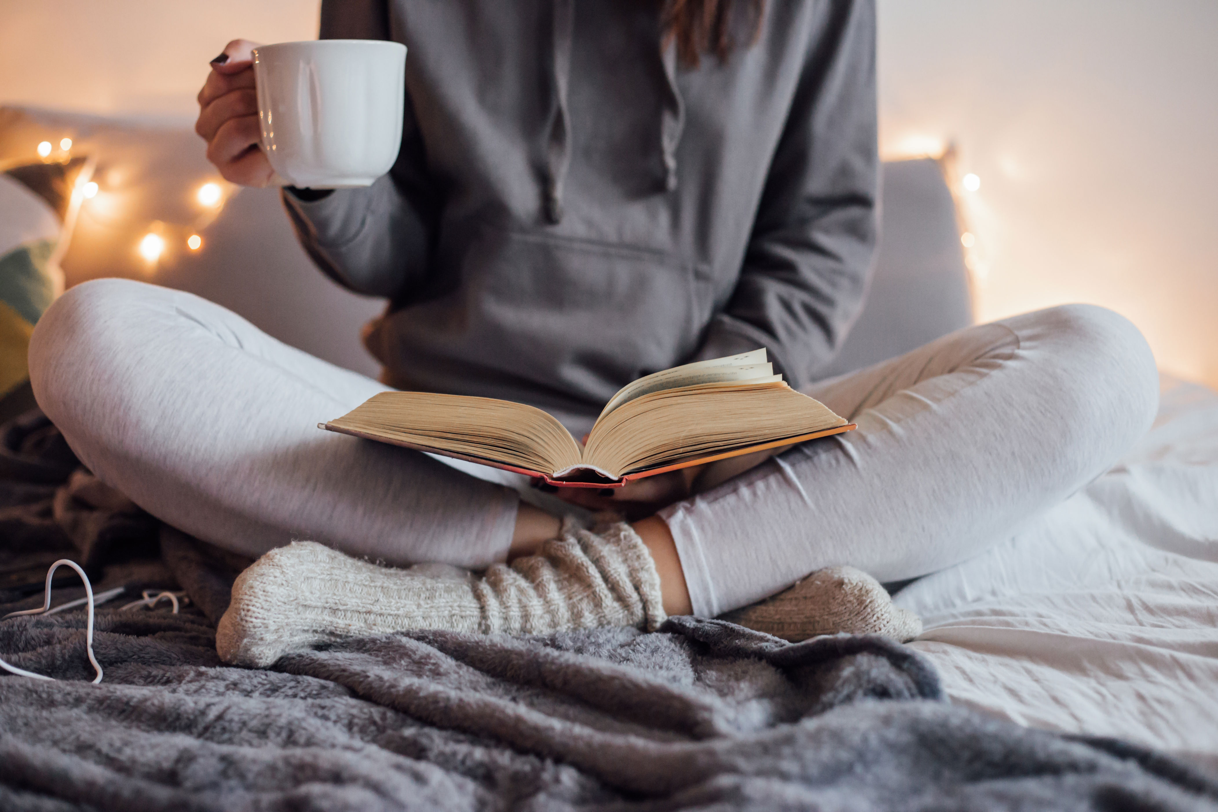 Person sitting cross-legged on a bed, holding a mug, and reading a book, suggesting a cozy, relaxed atmosphere