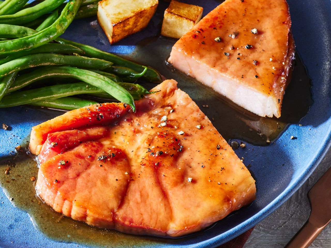A plate with a sliced brown sugar ham steak, topped with cracked black better and served with green beans and potatoes
