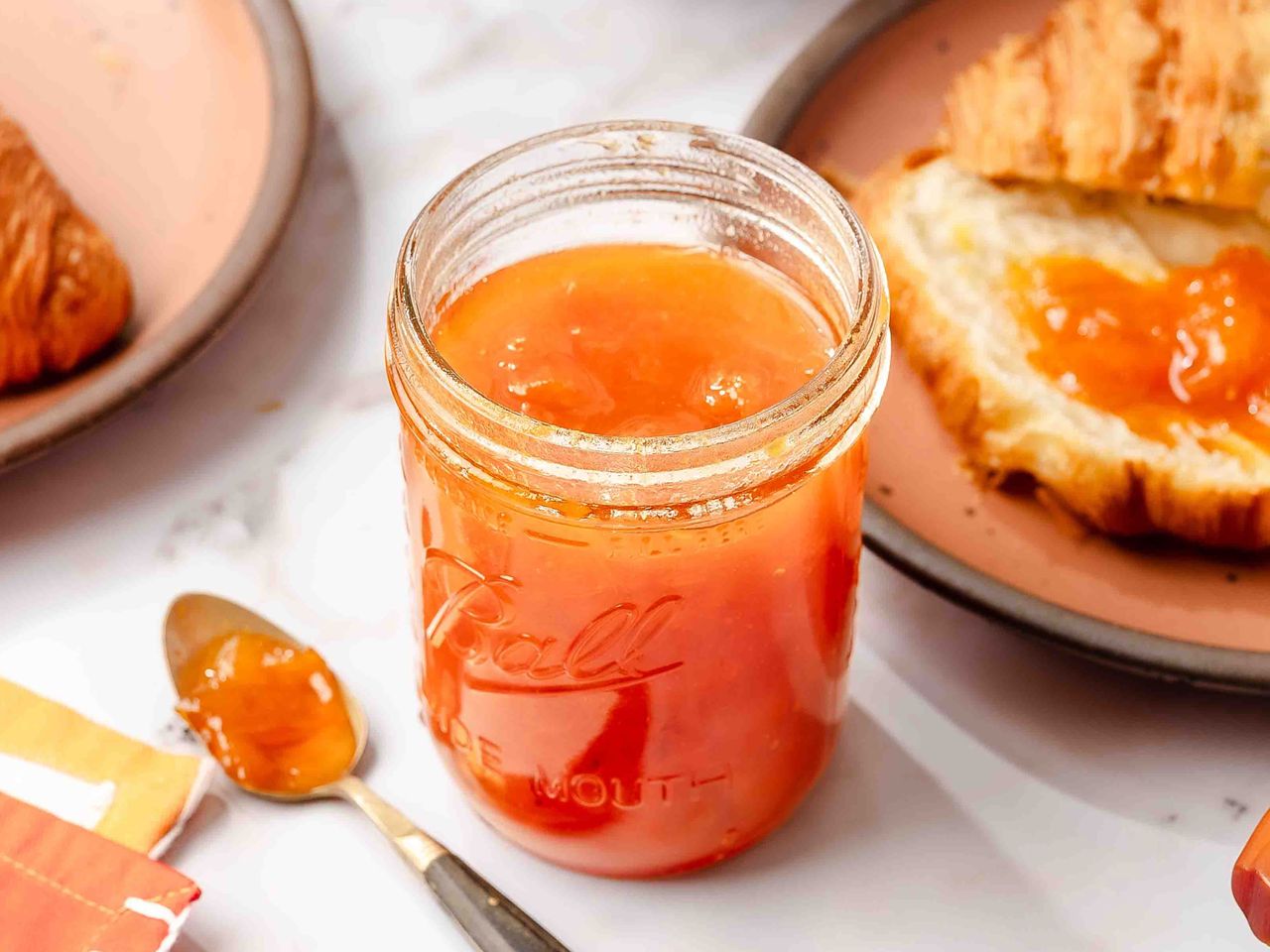 A jar of apricot jam with a spoon and croissants in the background