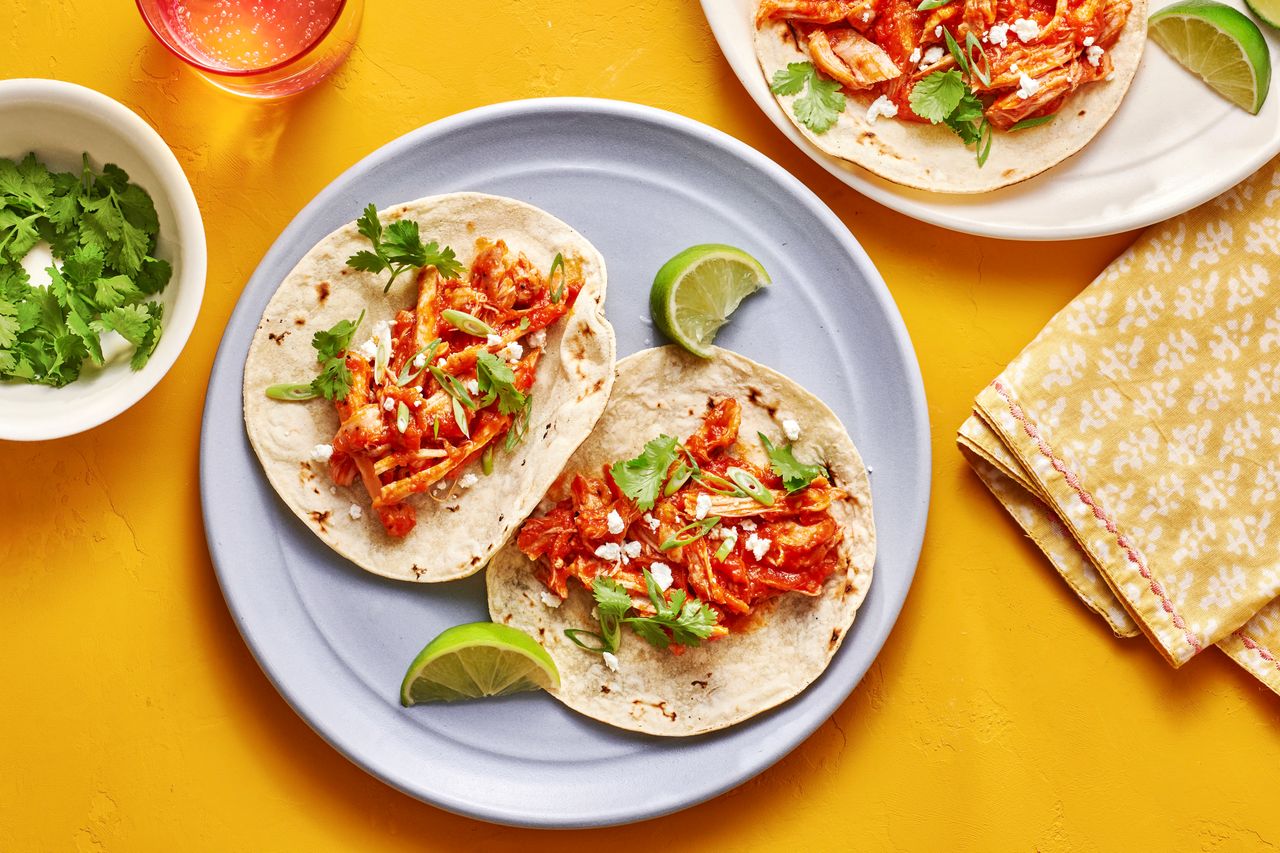 A plate with two chicken tinga tacos served with lime wedges, cilantro, and queso fresca