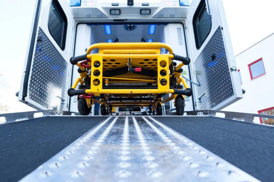 A yellow, specialized vehicle is being unloaded from a large vehicle through an open doorway, positioned on a paved ramp. The scene suggests an emergency medical transport operation.