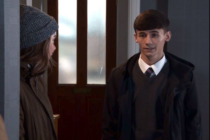 Two individuals are engaged in conversation in a corridor. One person, dressed in a beret, stands partially obscured. The other person, wearing a formal suit and tie, faces the observer. The background features a partially visible door with a glass panel.