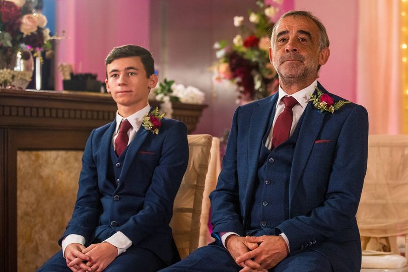Two individuals dressed in formal attire, including suits and ties, are seated together, possibly in a ceremonial setting, with a decorative background that includes a mantle and floral arrangements.