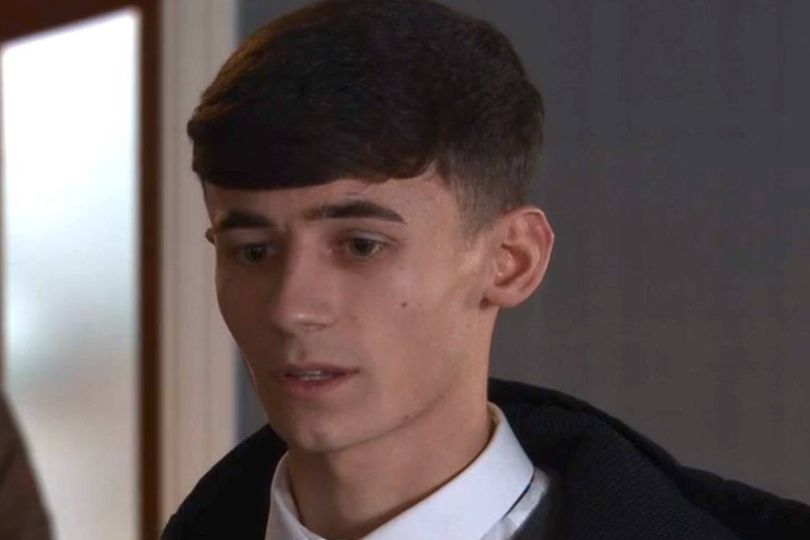 Two individuals, one dressed in a formal suit and tie, the other in a brown coat, stand in close proximity to an open door.