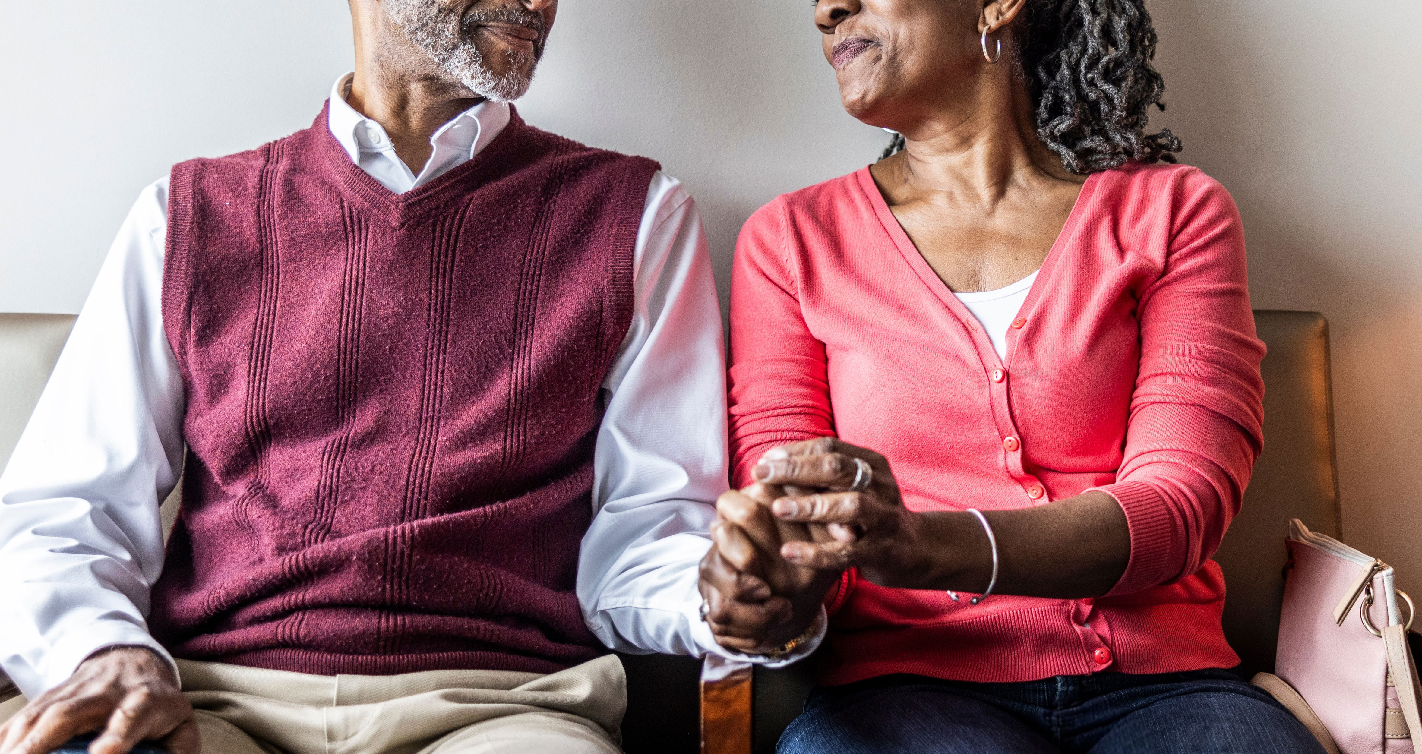An elderly couple sits on a couch holding hands and smiling at each other, next to a lamp on a side table