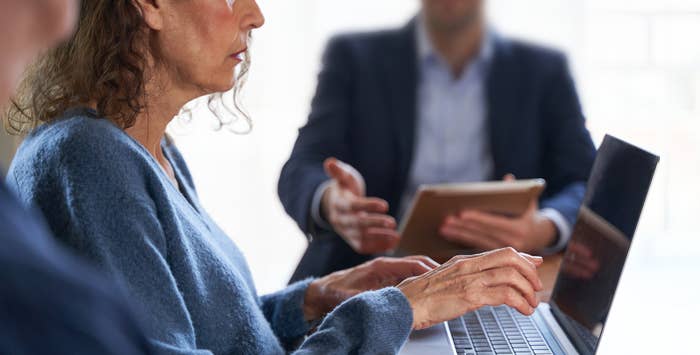 Woman typing on a laptop while a man in business attire speaks, holding a tablet, during a meeting