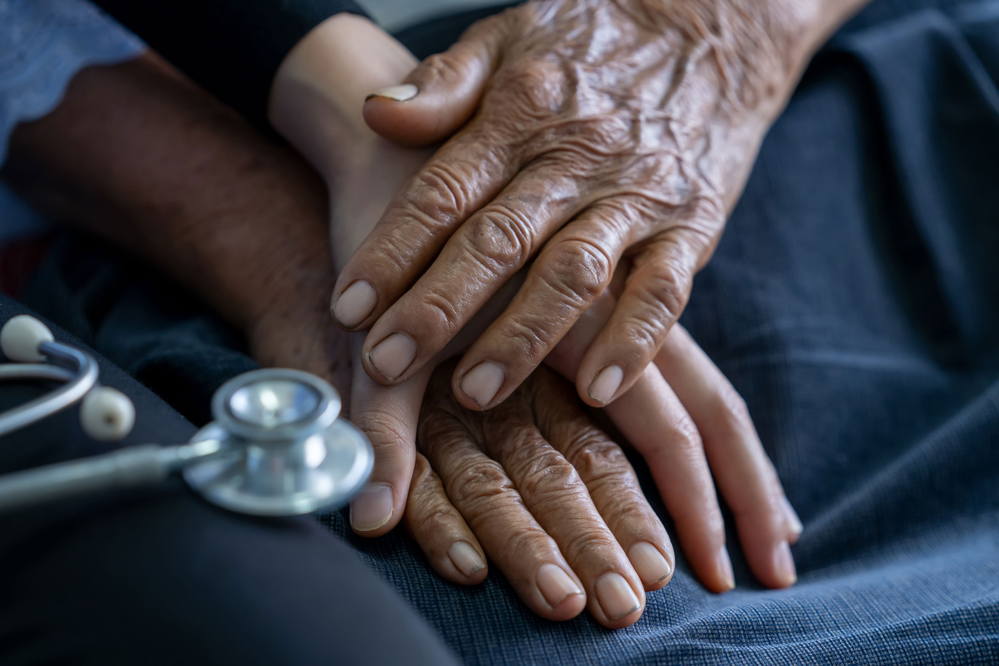 An elderly hand gently rests on a younger one, symbolizing care and support, with a stethoscope visible nearby
