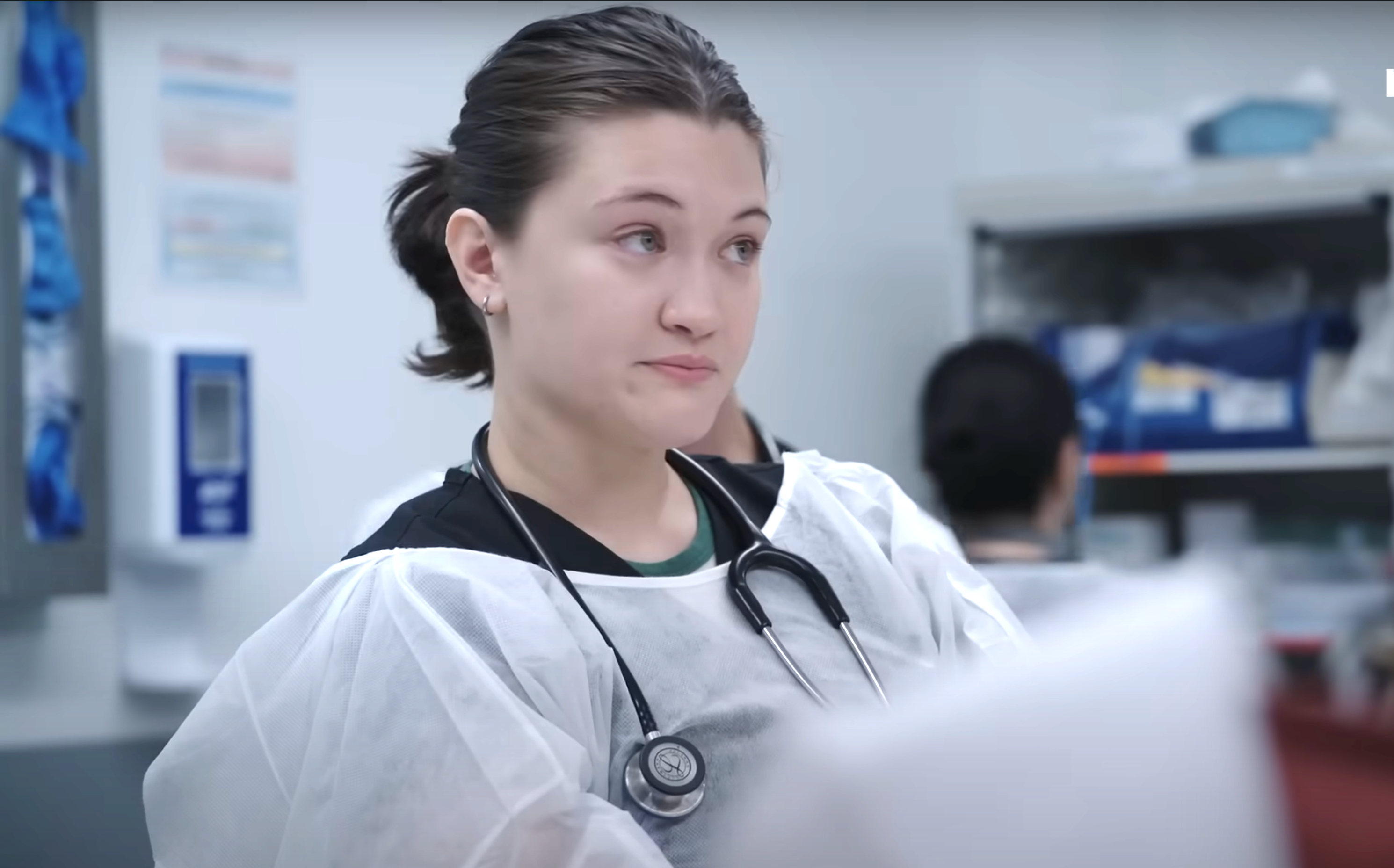 A healthcare professional wearing a stethoscope and protective gown looks attentive in a medical setting. Shelves with supplies are visible in the background