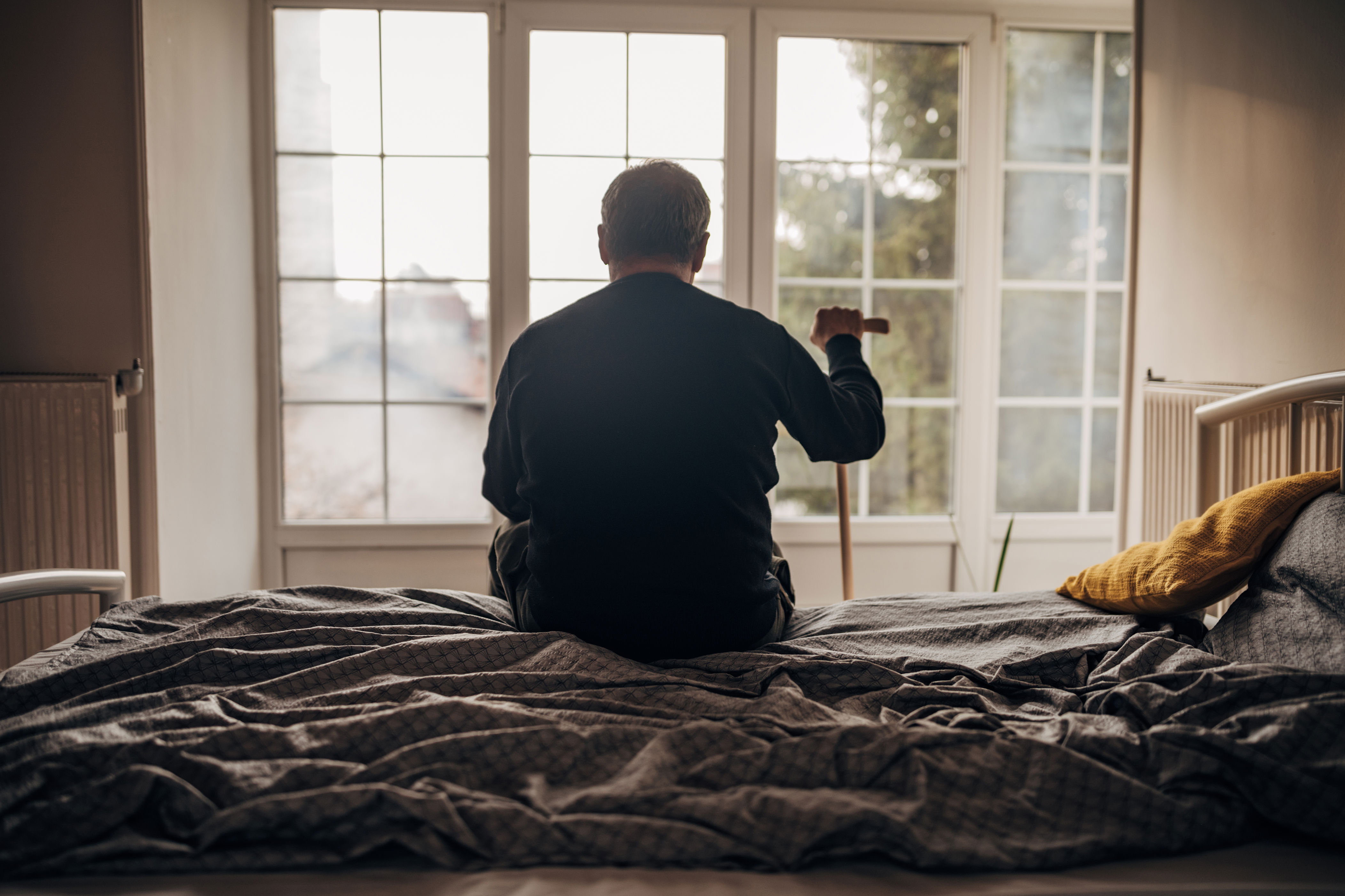 Person sitting on a bed, facing large windows, holding a walking stick. The room is softly lit, creating a contemplative atmosphere