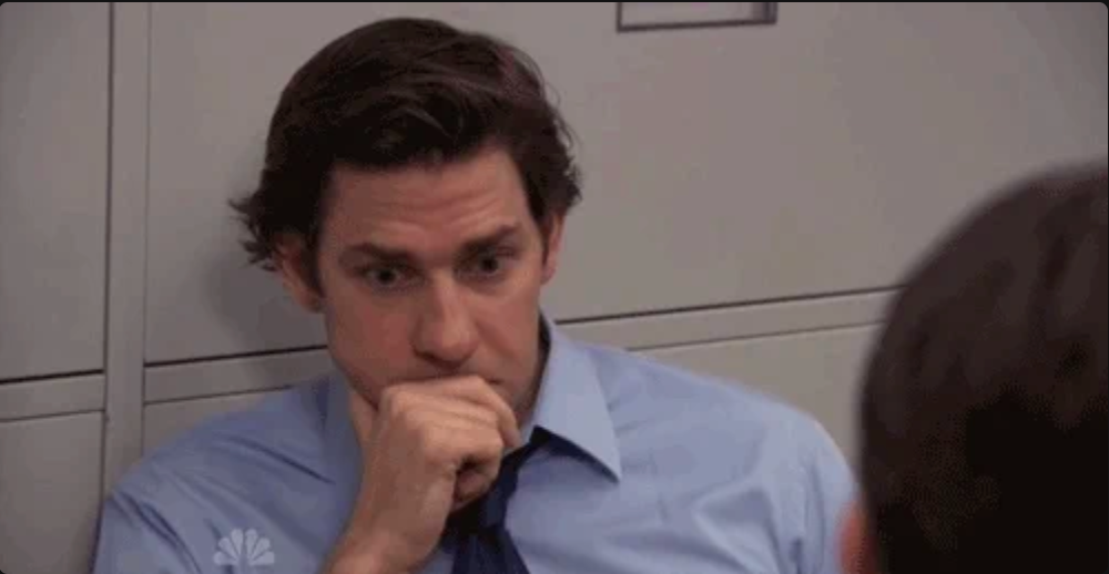 A person wearing a shirt and tie is seated indoors, looking contemplative, with their hand near their mouth