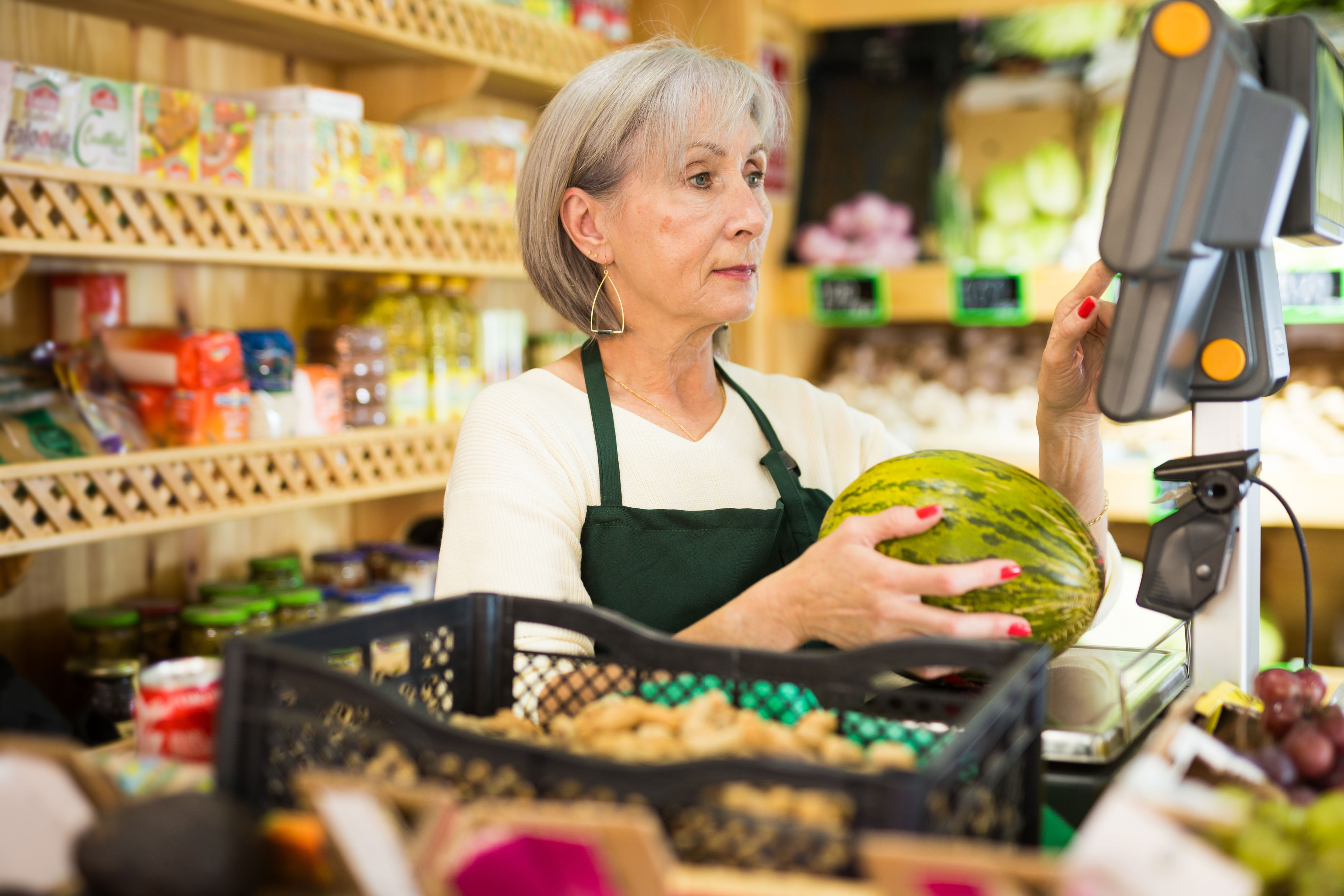 Woman in an apron holds a melon while operating a grocery store checkout. Shelves with jars and boxes are visible in the background