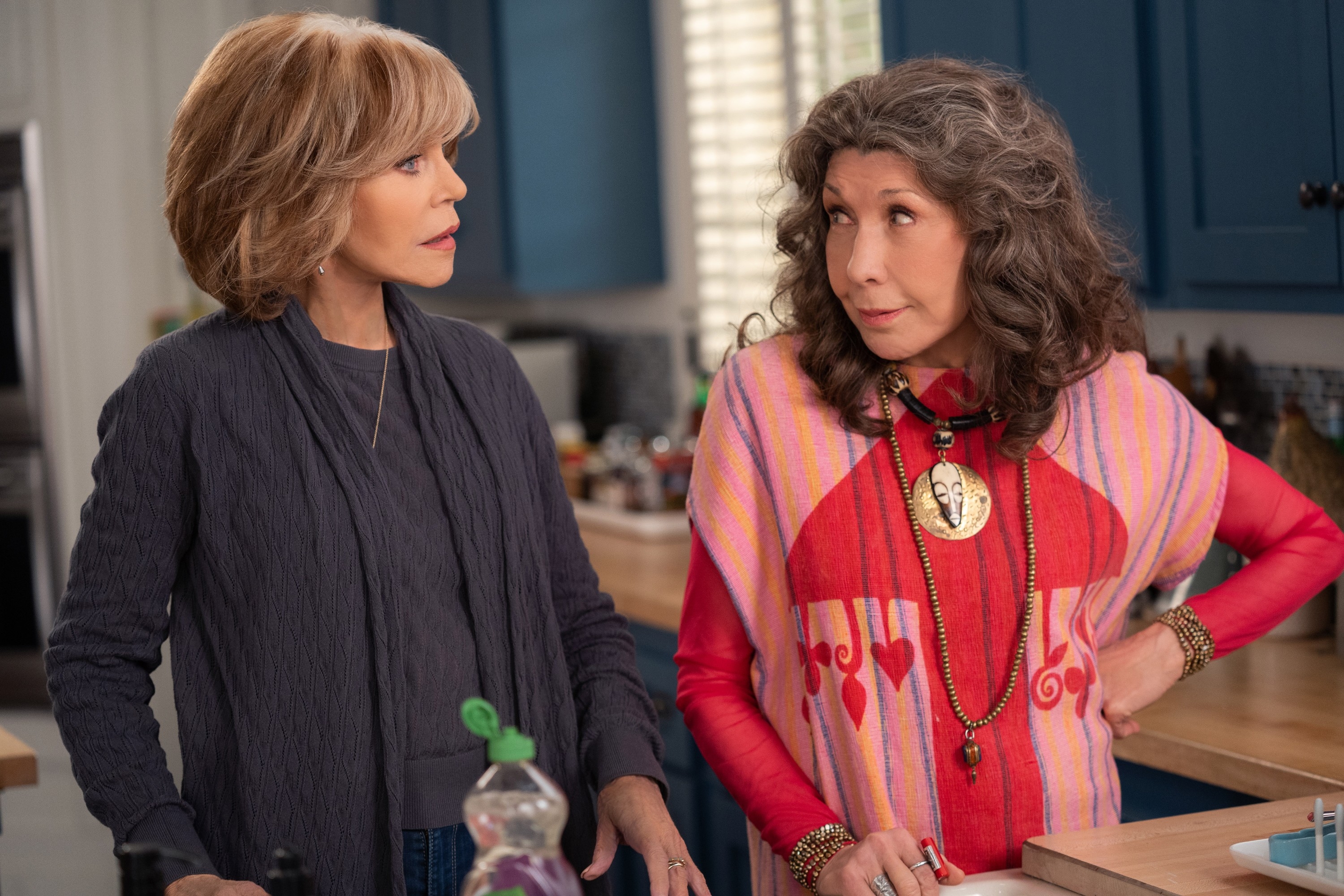 Two women in a kitchen, one in a flowy grey top and the other in a colorful, patterned shawl with bold jewelry, engaged in conversation
