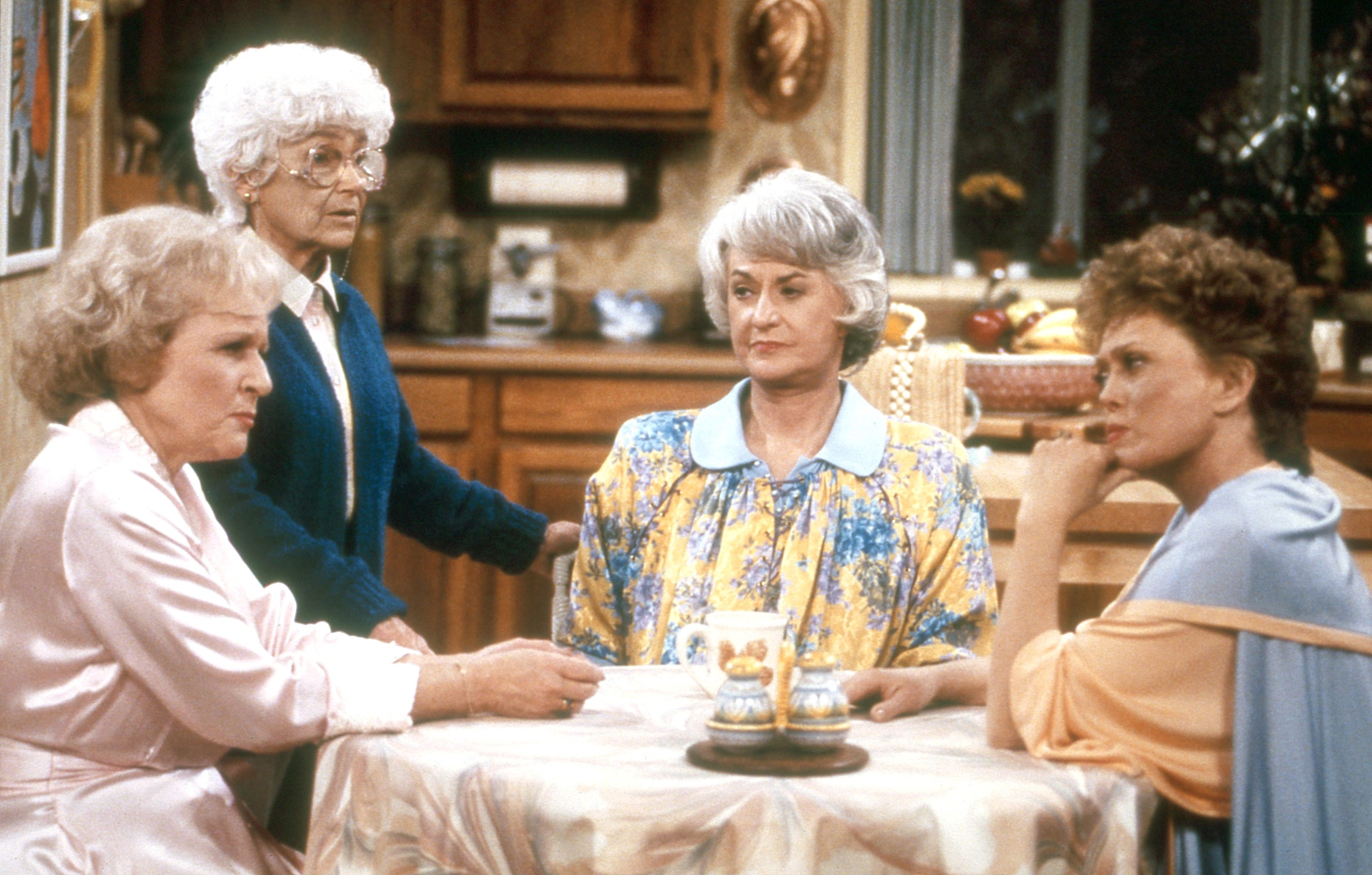 Four elderly women are seated at a kitchen table having a serious conversation