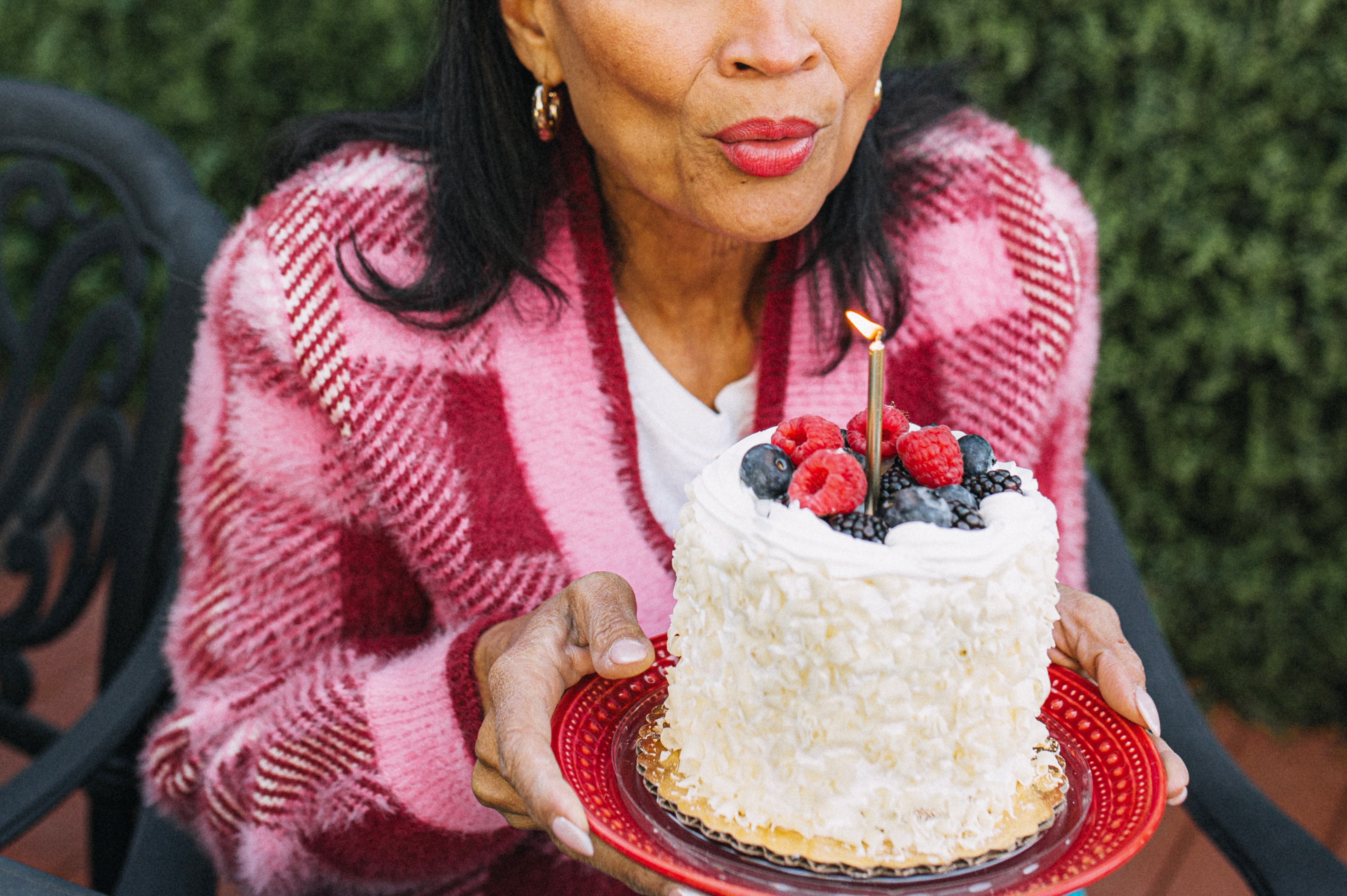 A person in a plaid jacket blowing out a candle on a small cake topped with berries, outdoors