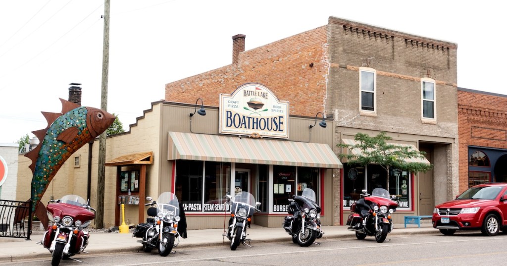 Motorcycles are parked in front of the Battle Lake Boathouse restaurant in downtown Battle Lake, Minnesota, next to a large mosaic fish sculpture.