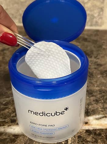 Hand using tweezers to pick a medicube Zero Pore Pad from an open container. The product is designed for skincare, featuring exfoliating ingredients