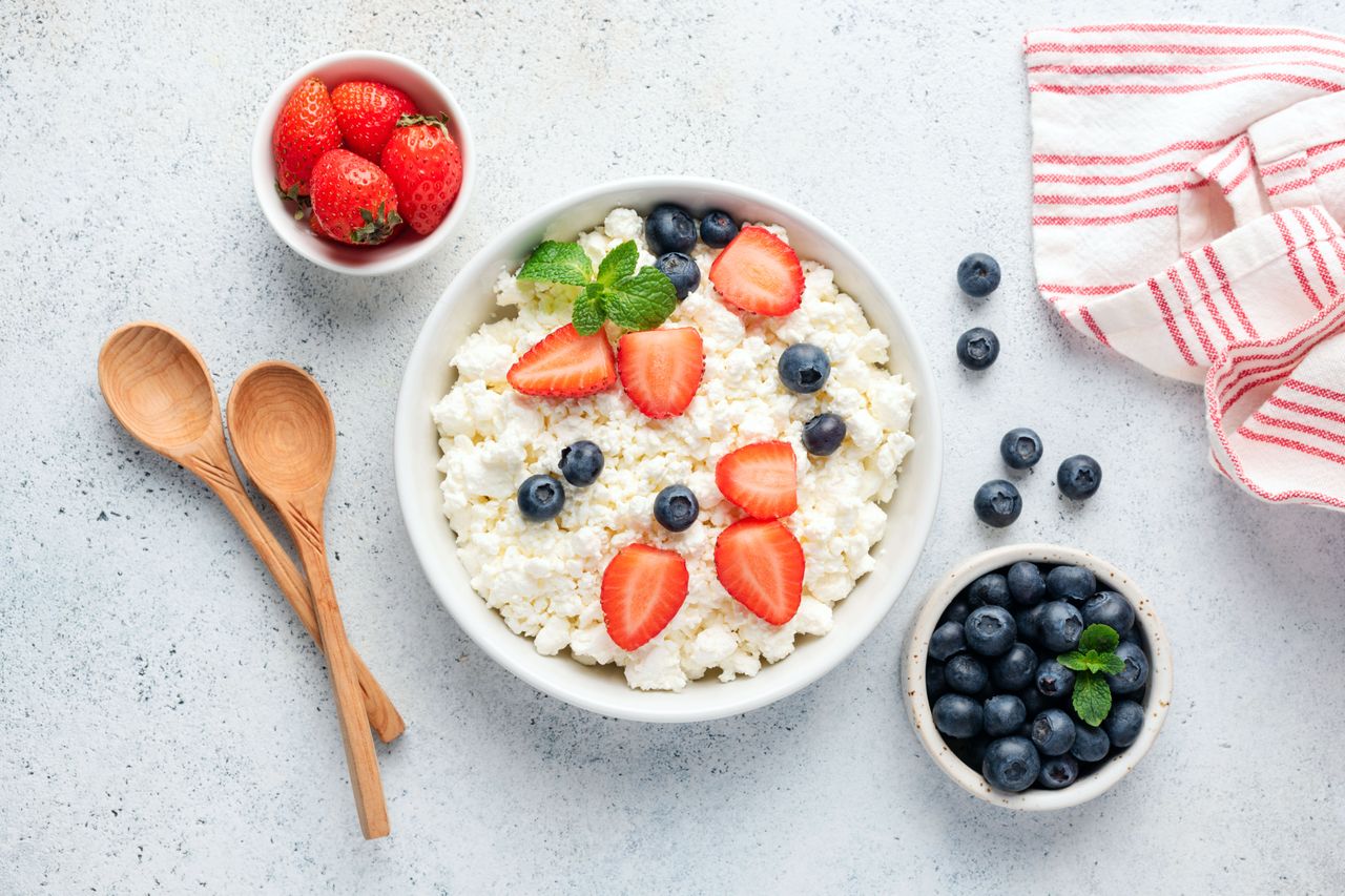 A bowl of cottage cheese topped with strawberries and blueberries surrounded by wooden spoons and small bowls of fruit