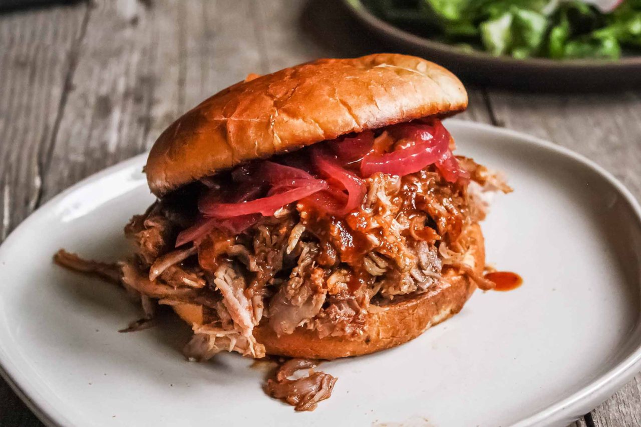 Pulled pork and pickled red onions in a toasted bun on a white plate, on a wooden table.