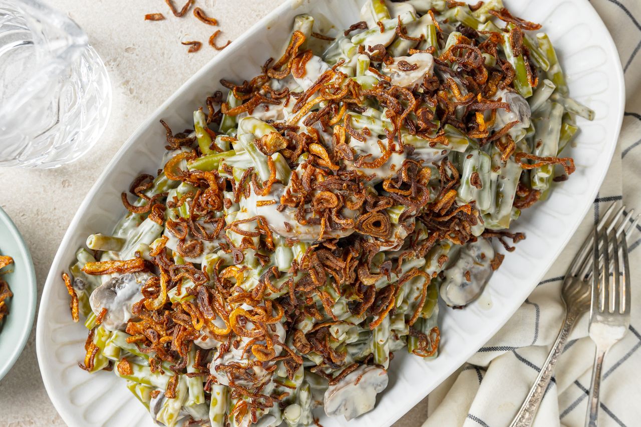 Slow Cooker Green Bean Casserole Topped with Crispy Shallots on a Platter Surrounded by Utensils, a Kitchen Towel, and a Glass of Water