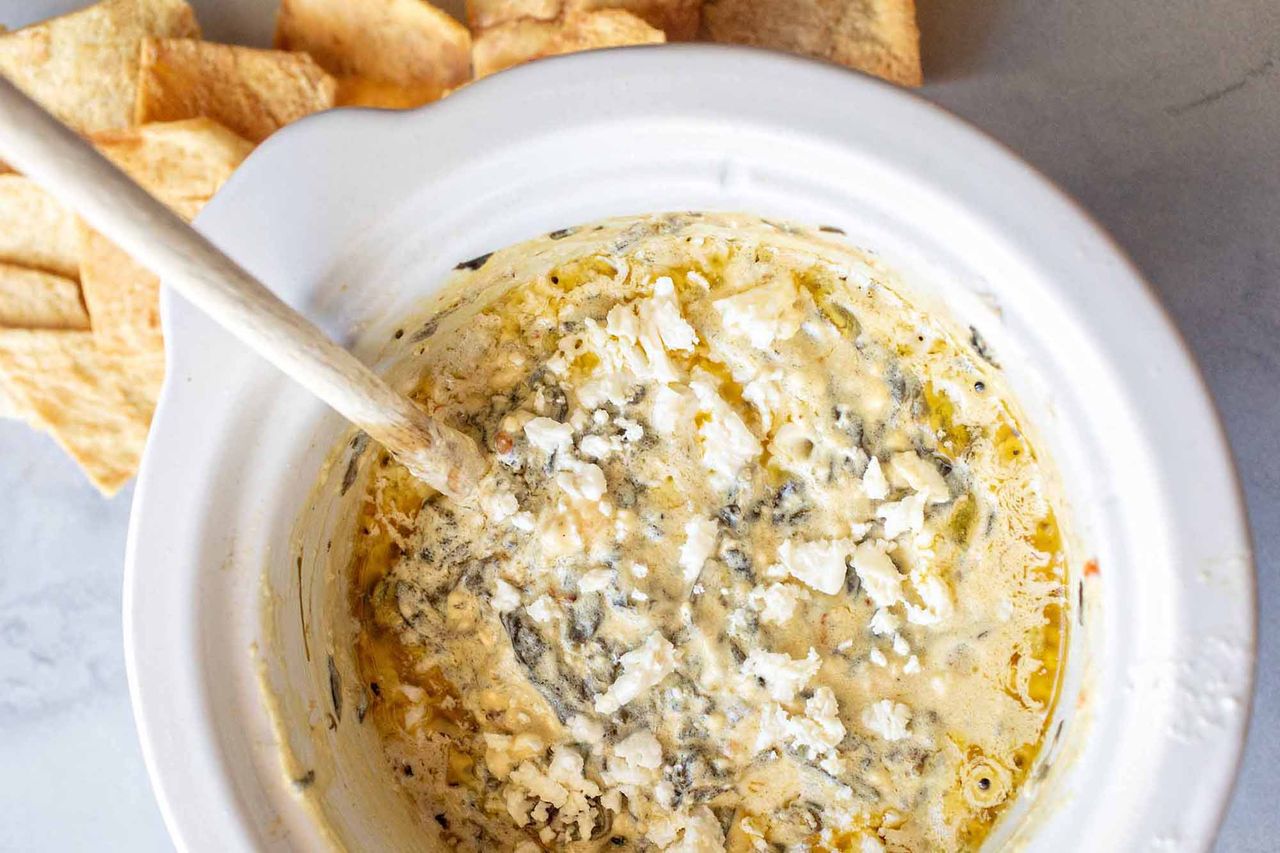 A white bowl of spinach dip with crumbled feta on top, next to some pita chips on a white marbled background.