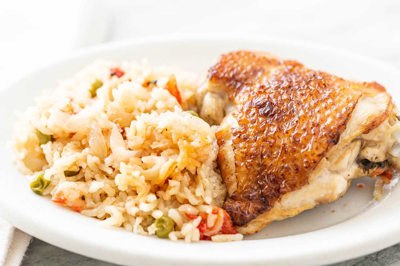A piece of cooked chicken next to a mound of rice and vegetables on a white plate.