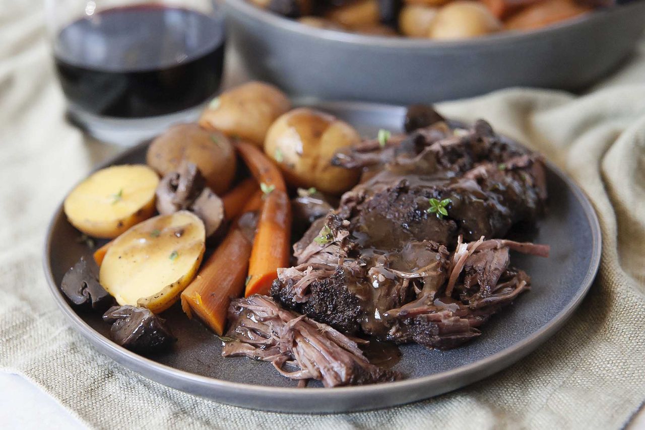 A plate of pot roast with potatoes and carrots on a dark grey plate, on an off-white fabric background.
