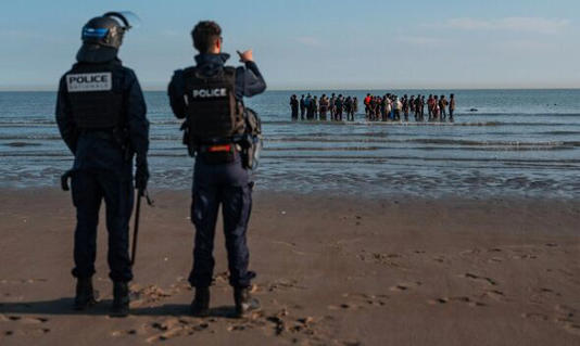 Police officers look on as migrants wade in the sea to try and board a migrant dinghy to sail into the English Channel on June 17, 2025 in Gravelines,