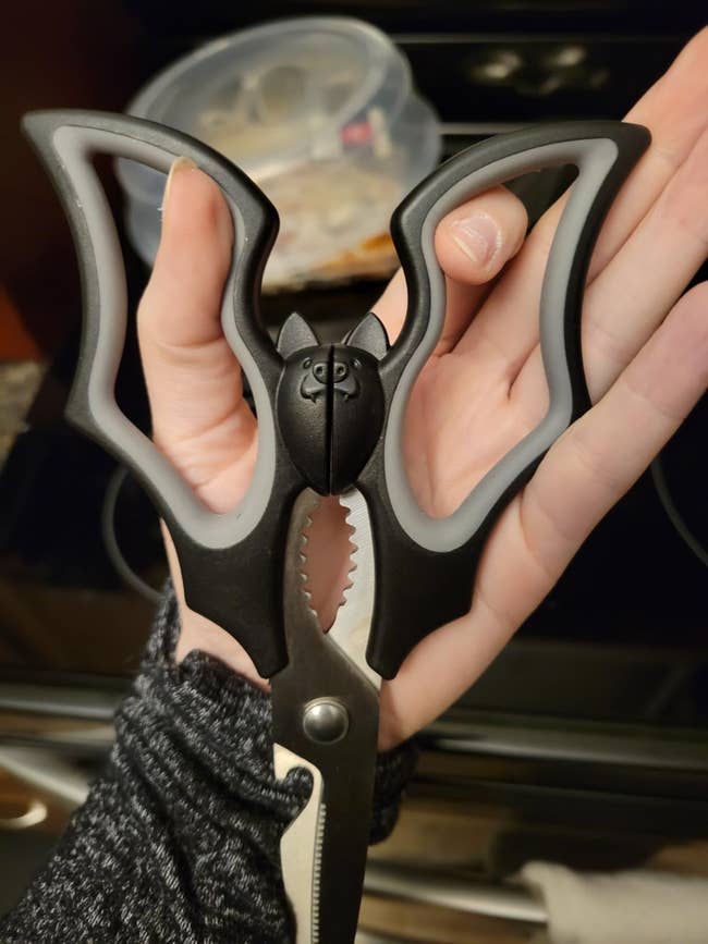 Person holding novelty kitchen scissors with a black bat-shaped handle, resembling wings, suitable for themed cooking or gifting