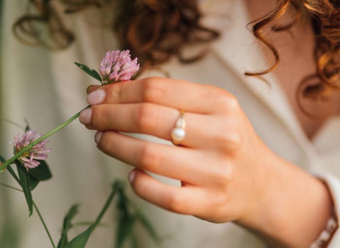 Person with curly hair holding a small flower, wearing a pearl ring and a white shirt, in a peaceful outdoor setting
