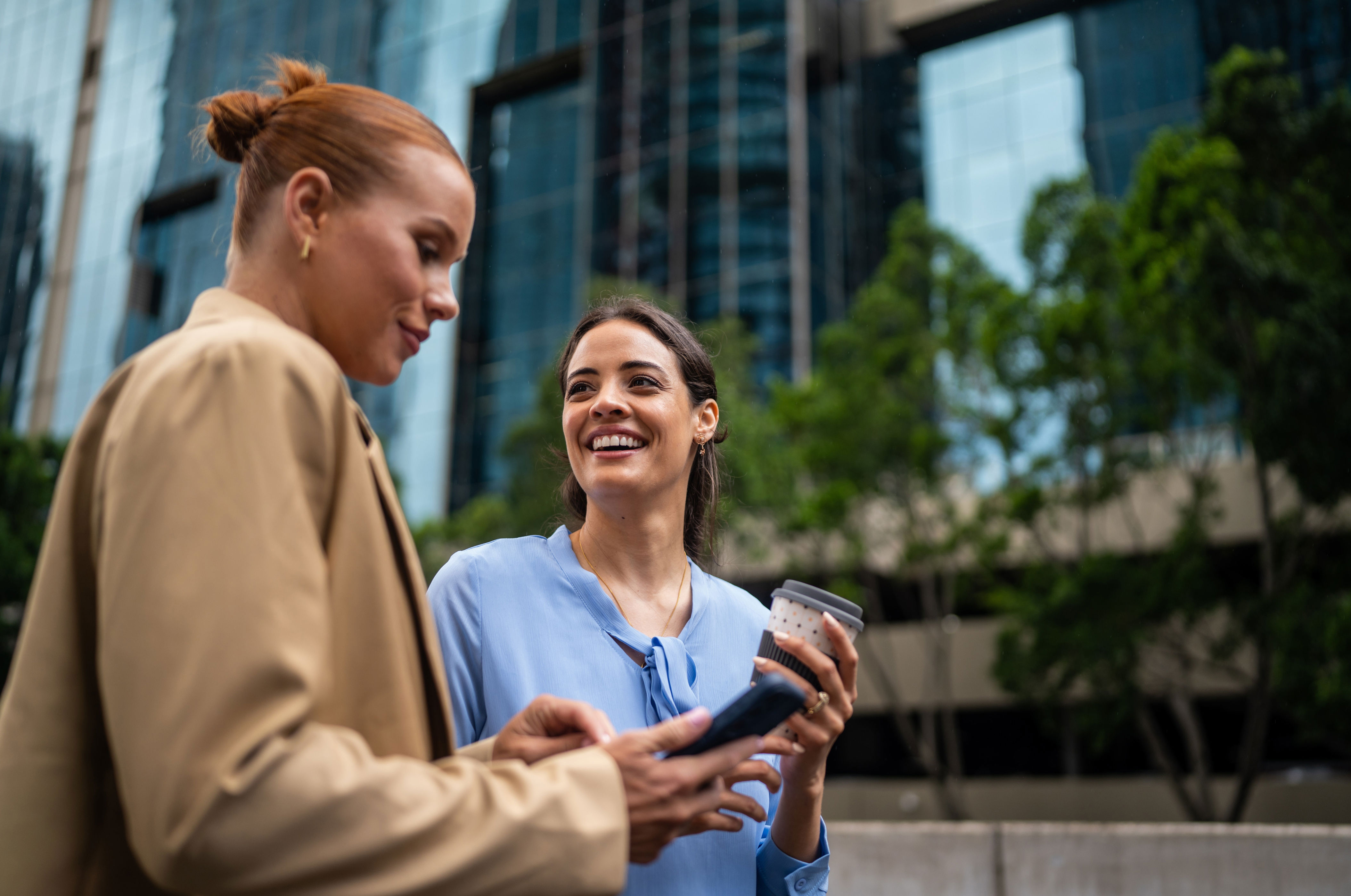 Two women smiling outdoors, one holding a phone and the other a cup. They're dressed in casual business attire, with city buildings in the background