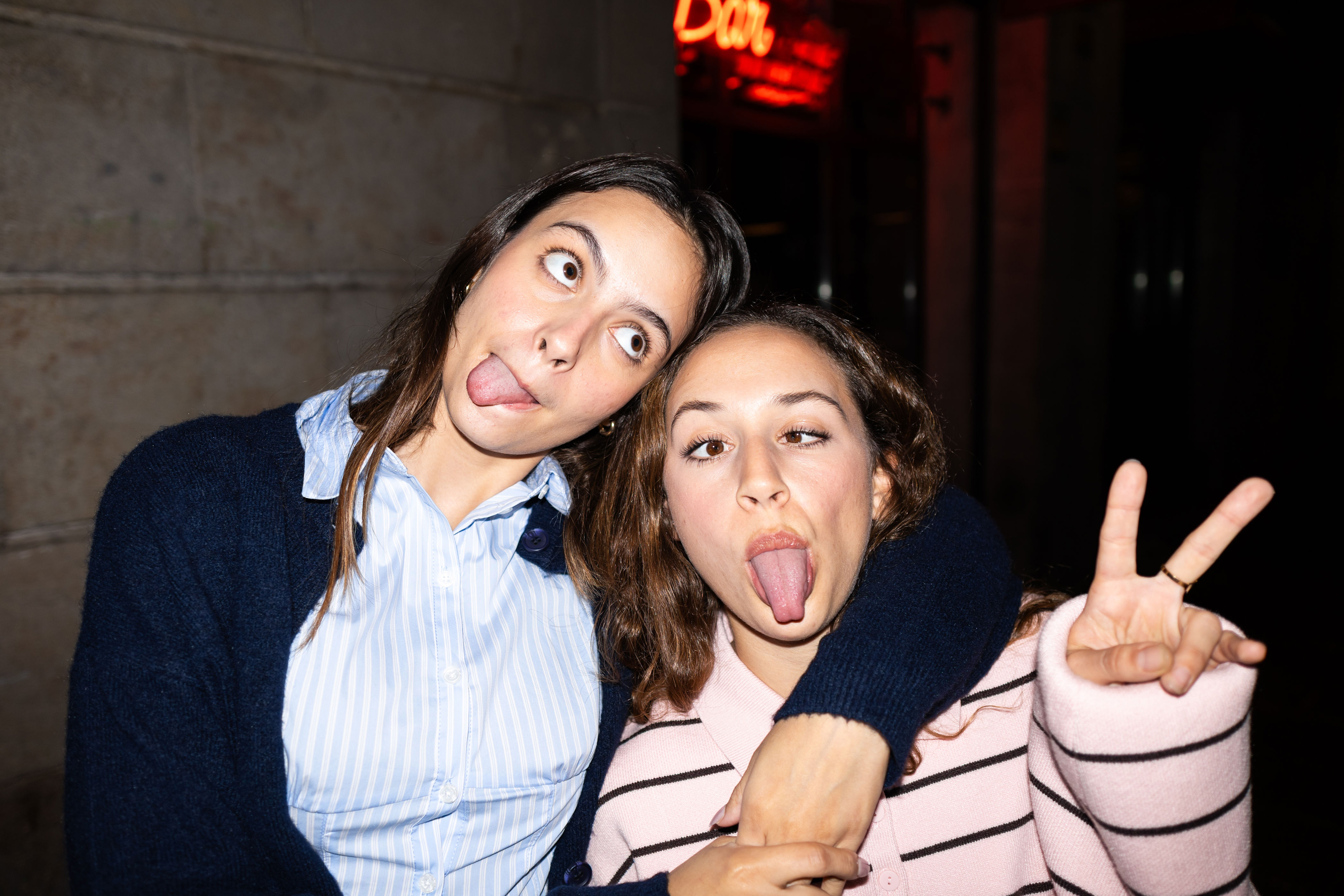 Two people make silly faces with tongues out. One gives a peace sign while the other wraps an arm around their shoulder. Casual, playful moment