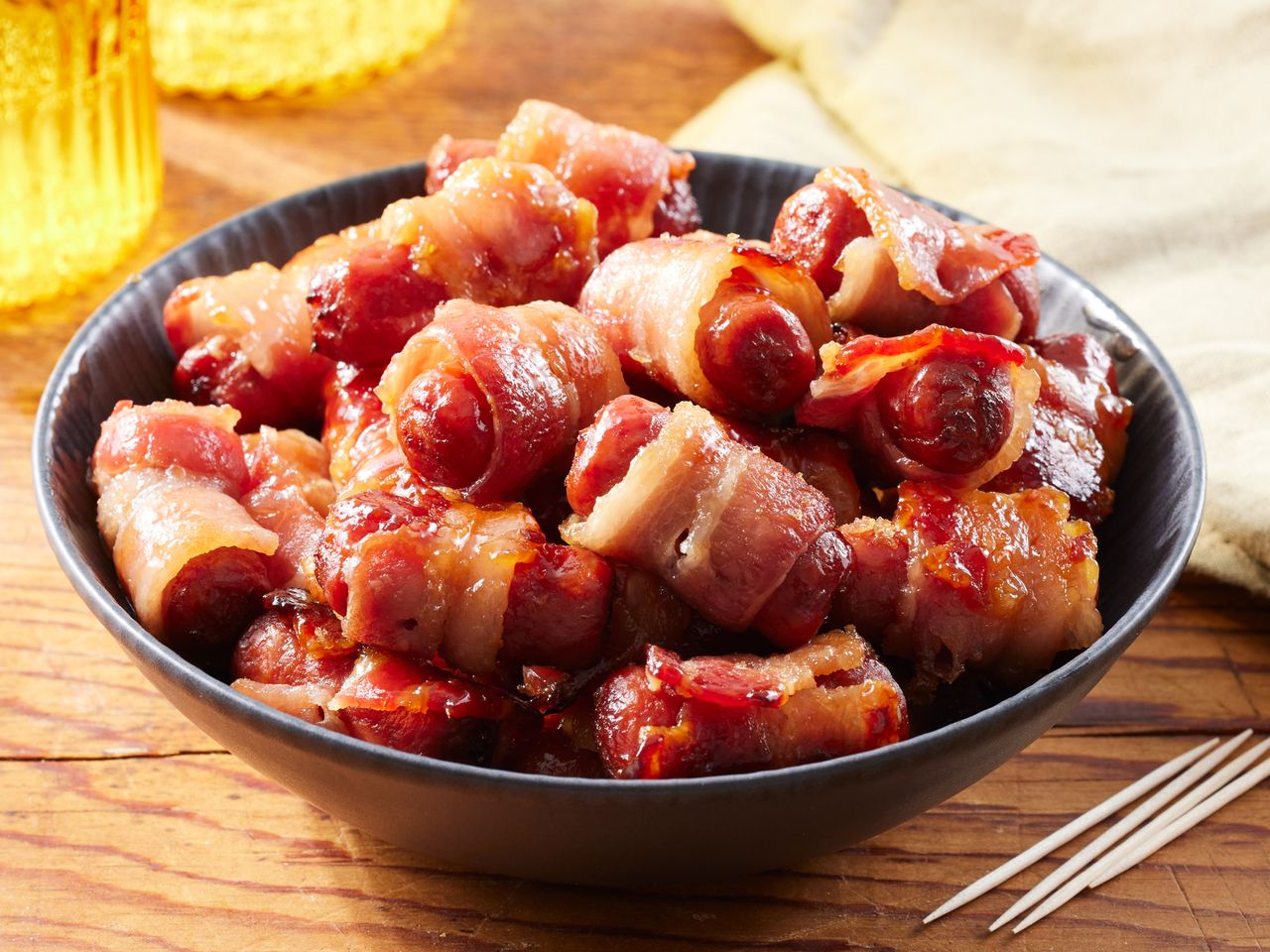 A bowl of brown sugar smokies (smokie sausages wrapped in bacon) served with toothpicks on the side