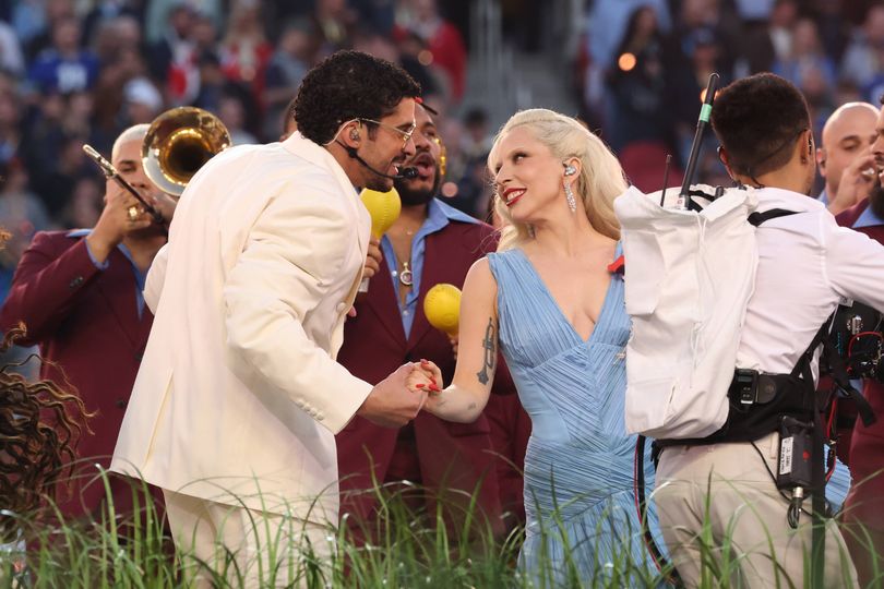 An individual in a white suit and another in a blue dress are standing on a grassy field, shaking hands, with musicians and other individuals in the background.