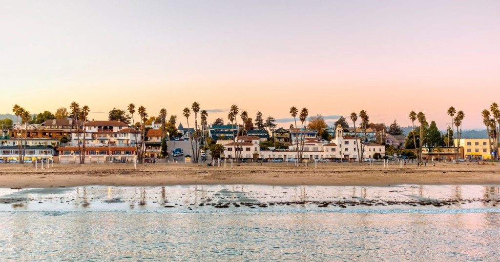 The sky turns pink and gold over a quaint beachside town with palm trees bordering the houses.