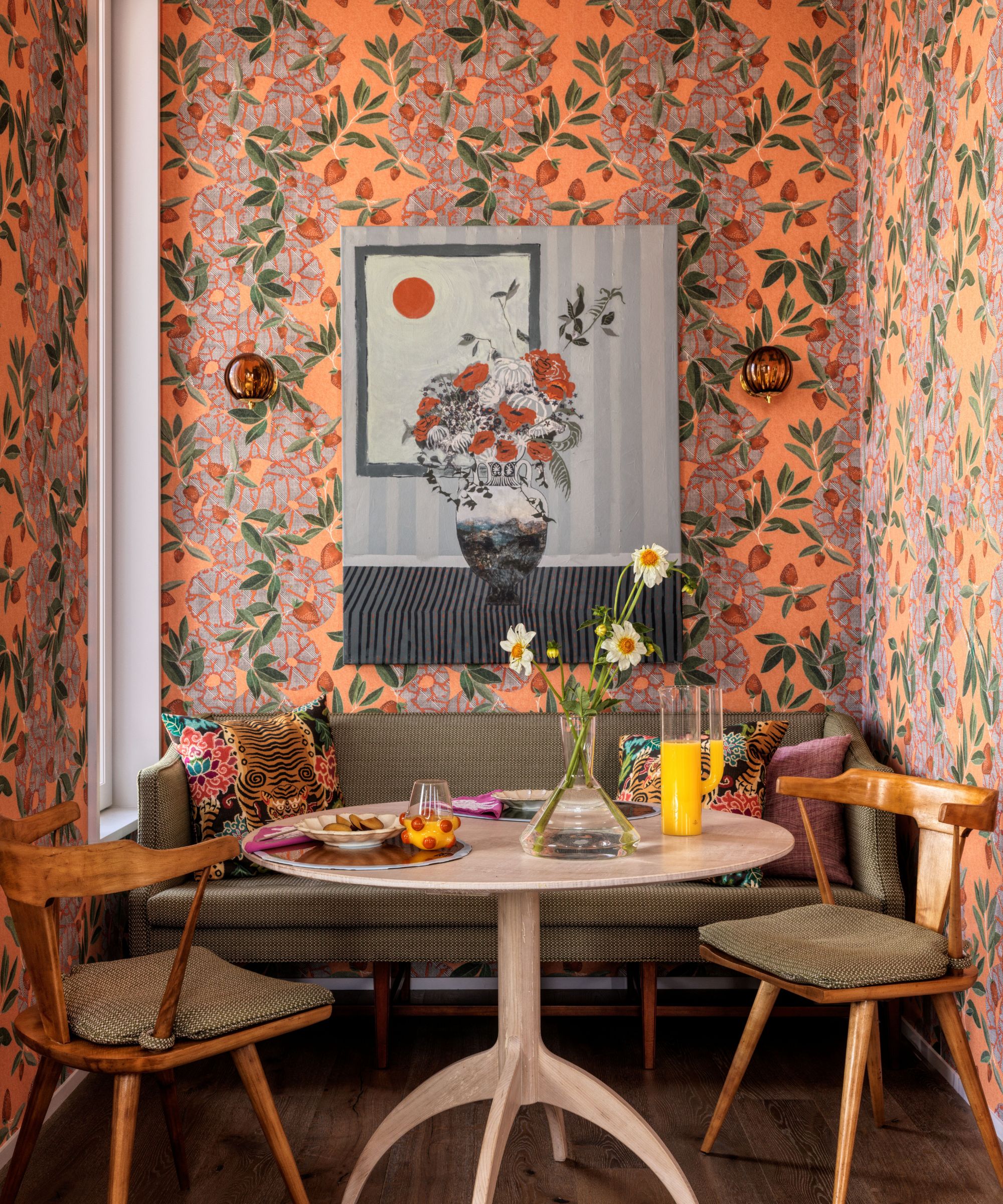 Colorful breakfast nook with orange floral wallpaper, bench, round wooden table, two vintage dining chairs and a painting of a floral arrangement