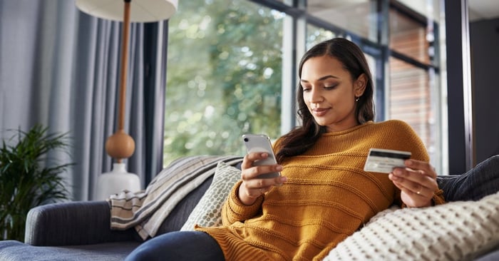 A woman sitting comfortably on her couch looking at her phone and holding a credit card.