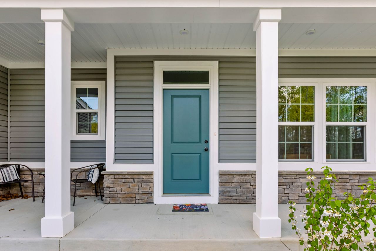 A front porch with a teal door flanked by two windows and decorative chairs on either side