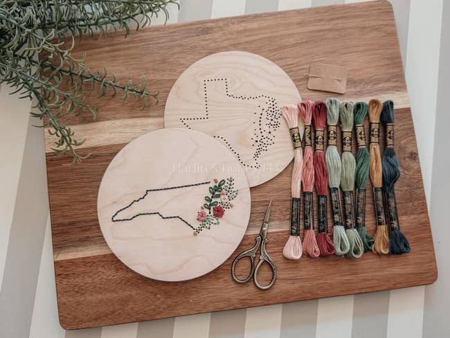 Embroidery kit with wooden hoops, floral design, and skeins of colorful thread on a wooden board. Scissors beside the kit